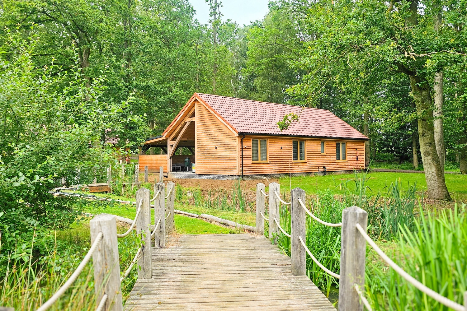 Ground-level hot tub lodge at Hay’s Wood Retreat in Norfolk, UK.  A charming wooden bridge and winding path surrounded by trees and greenery shows the lodge in the distance. Designed for easy access, these self-catering lodges offer guests a tranquil countryside retreat with private hot tubs, blending comfort with nature in a peaceful woodland setting.